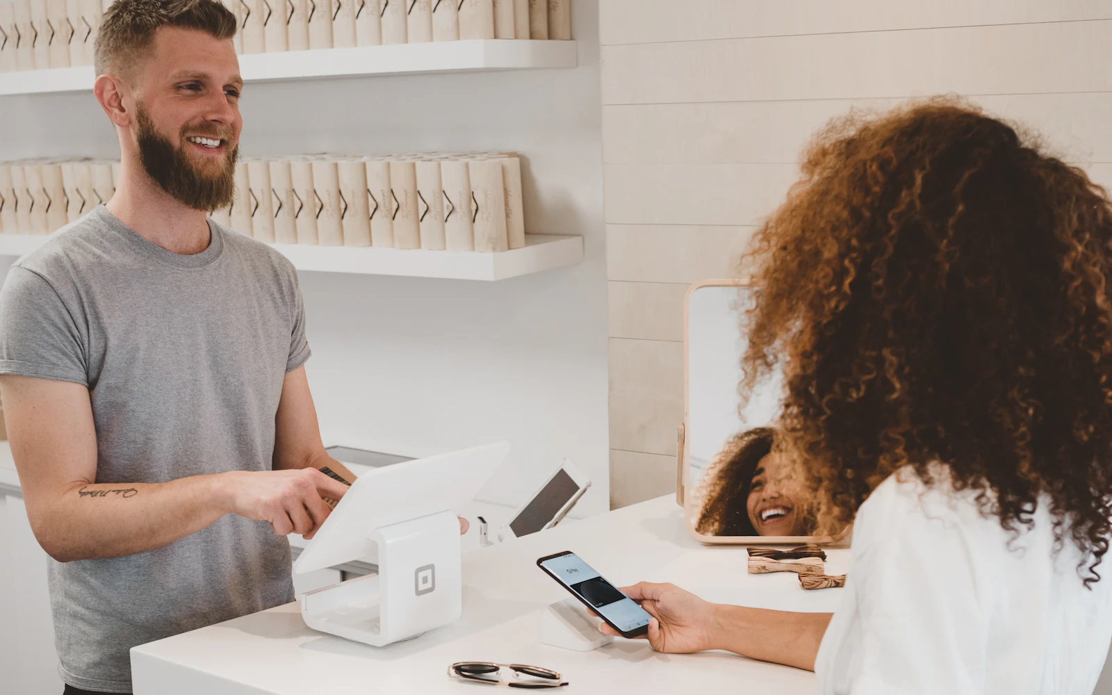 A customer holding their phone to a card reader at a bright shop counter while the shopkeeper works the till