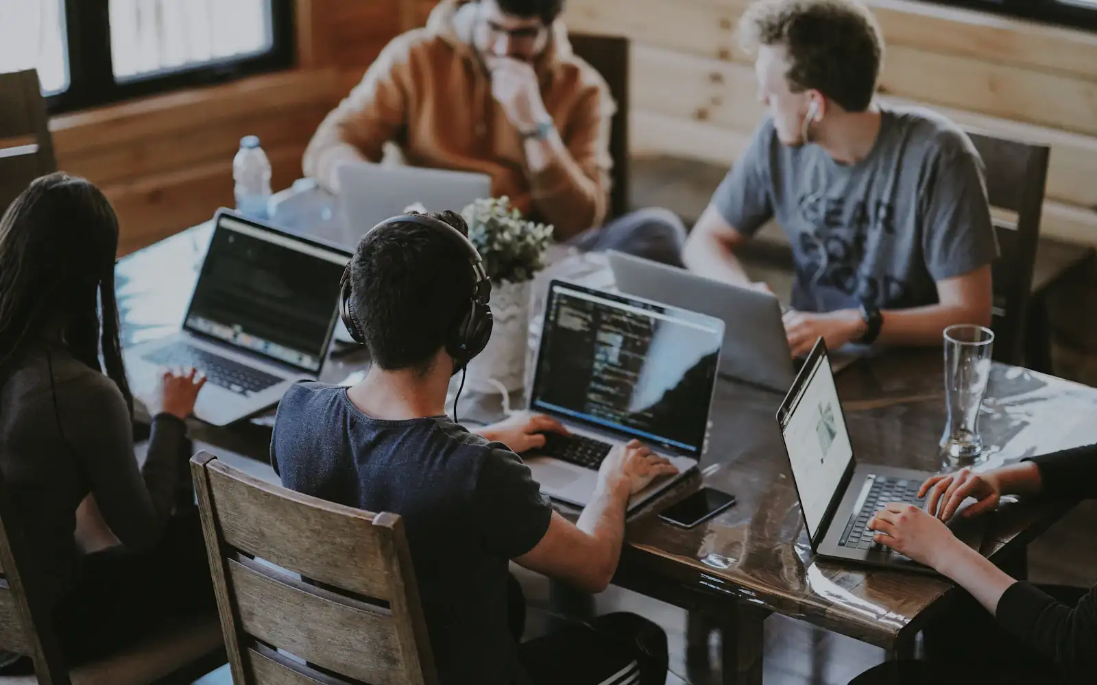 Five people around a wooden table, each working on a laptop, one of them in headphones