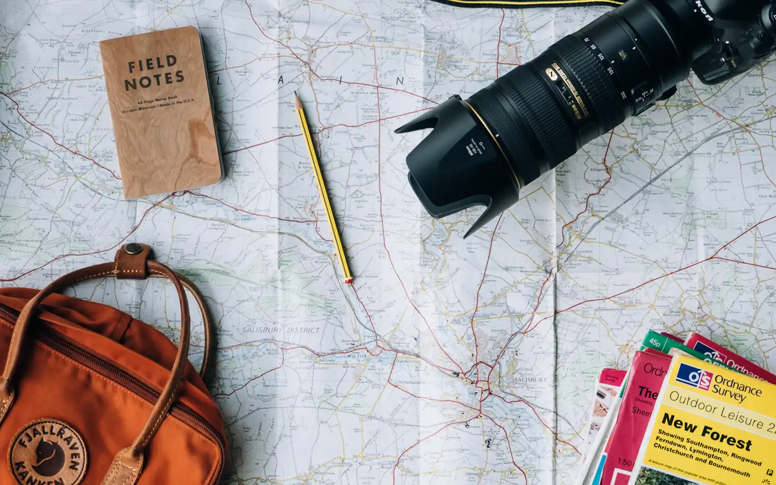 A paper Ordnance Survey map with a camera and telephoto lens, a Field Notes pad, a pencil and an orange backpack laid on top