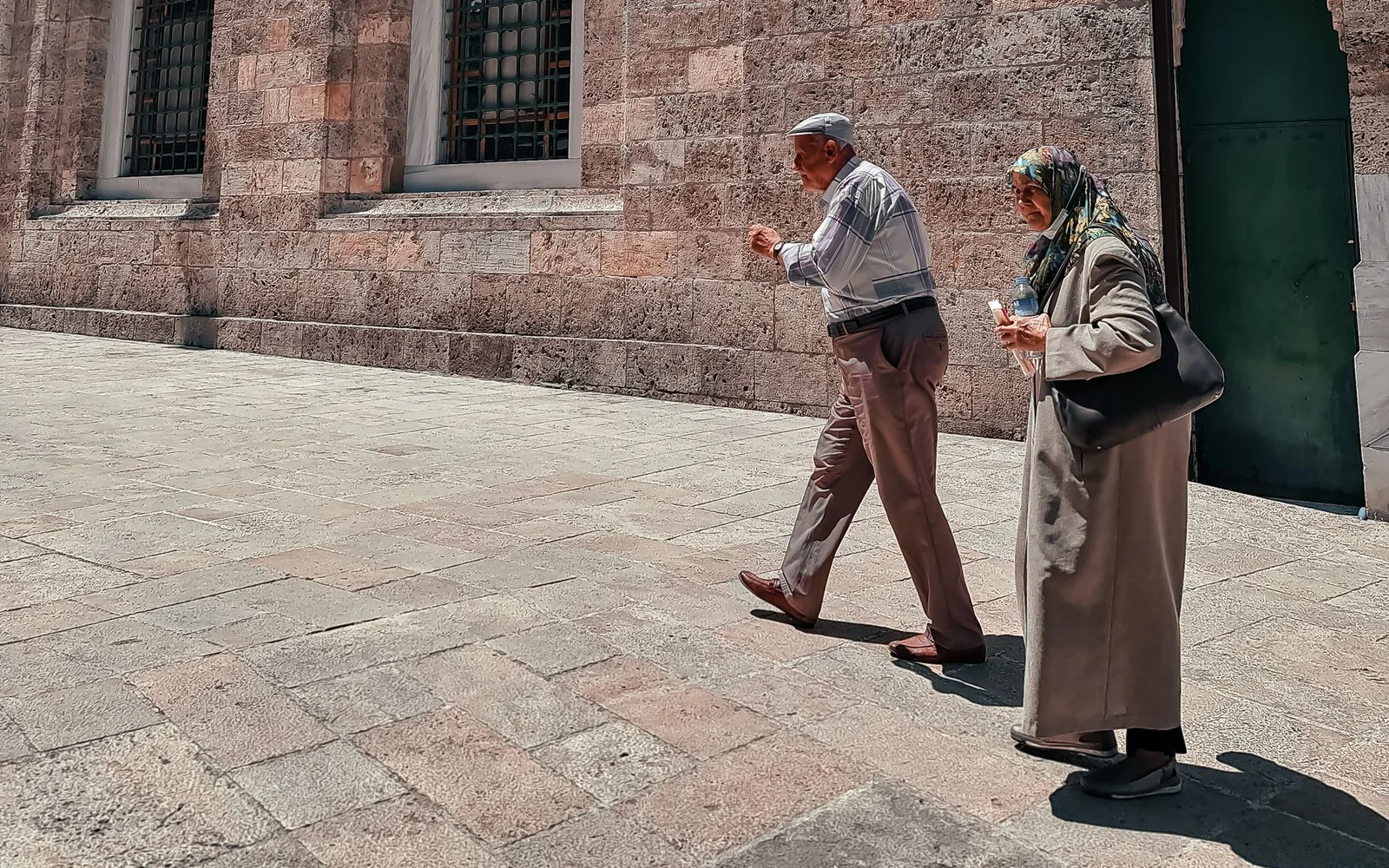 Two older adults walking beside a stone building on a paved street